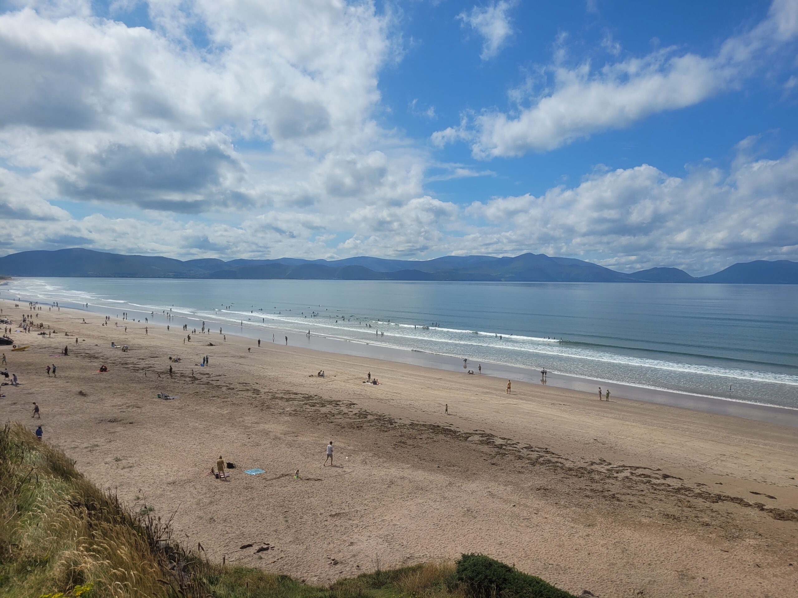 Expansive sandy beach at Inch Beach in County Kerry with people walking and relaxing, gentle waves, partly cloudy sky, and mountains in the background.
