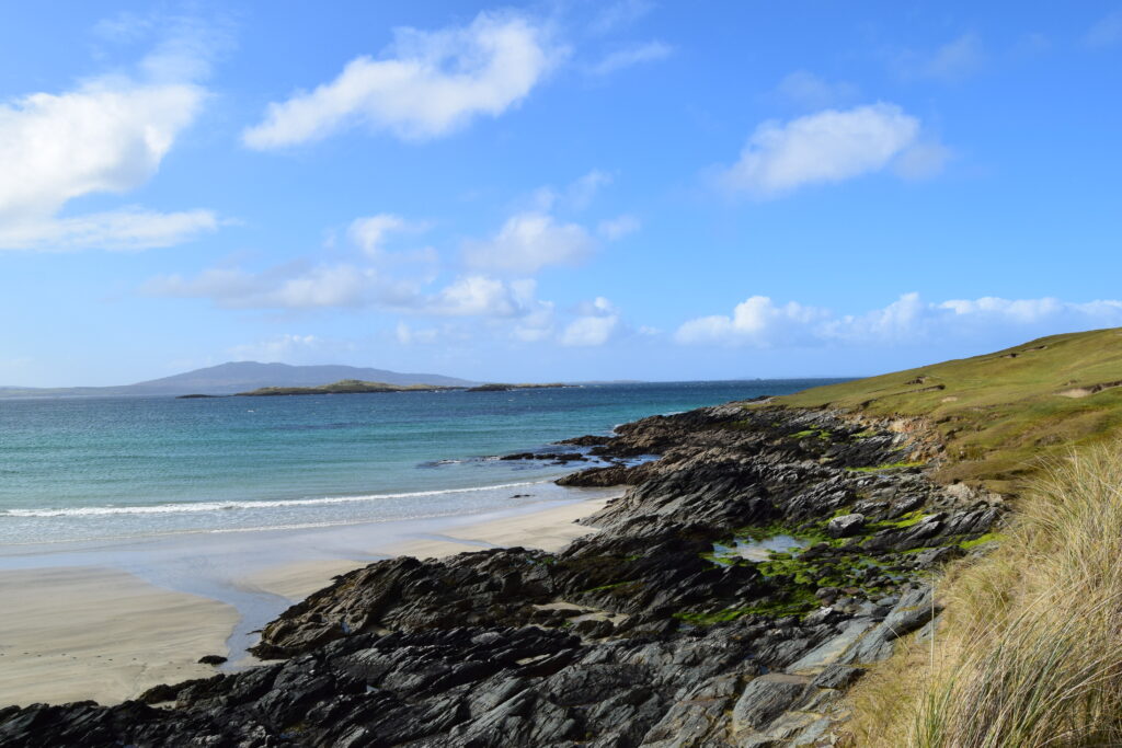 A coastal landscape featuring a sandy beach, rocky shoreline, and grassy hills. The ocean appears calm with gentle waves, and distant islands or landmasses are visible on the horizon. The sky is mostly clear with scattered clouds.
