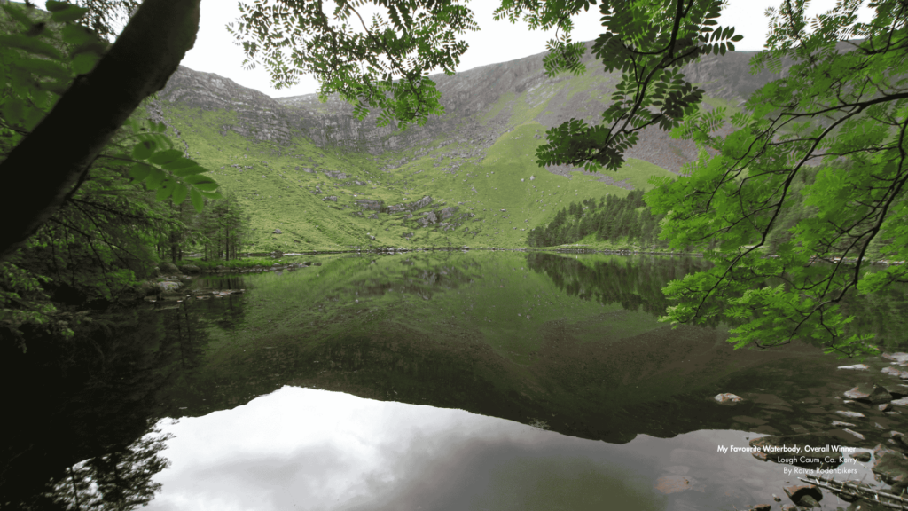 A tranquil landscape of Lough Caum in County Kerry, Ireland, showing a calm lake surrounded by green hills and trees, with their reflections mirrored on the water. Foreground branches with green leaves frame the scene. Text in the bottom right corner reads: 'My Favourite Waterbody, Overall Winner Lough Caum, Co. Kerry By Raivis Rodenbikers
