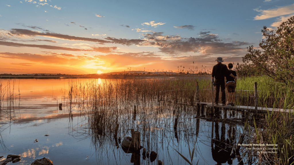 A peaceful sunset scene over the River Shannon in County Longford, Ireland. Two people stand on a wooden dock, gazing at the partly cloudy sky with orange and blue hues reflected on the calm lake. Tall reeds rise from the water in the foreground. Text in the bottom right corner reads: 'My Favourite Waterbody, Runner-up River Shannon, Co. Longford By Brigid Mullooly.'