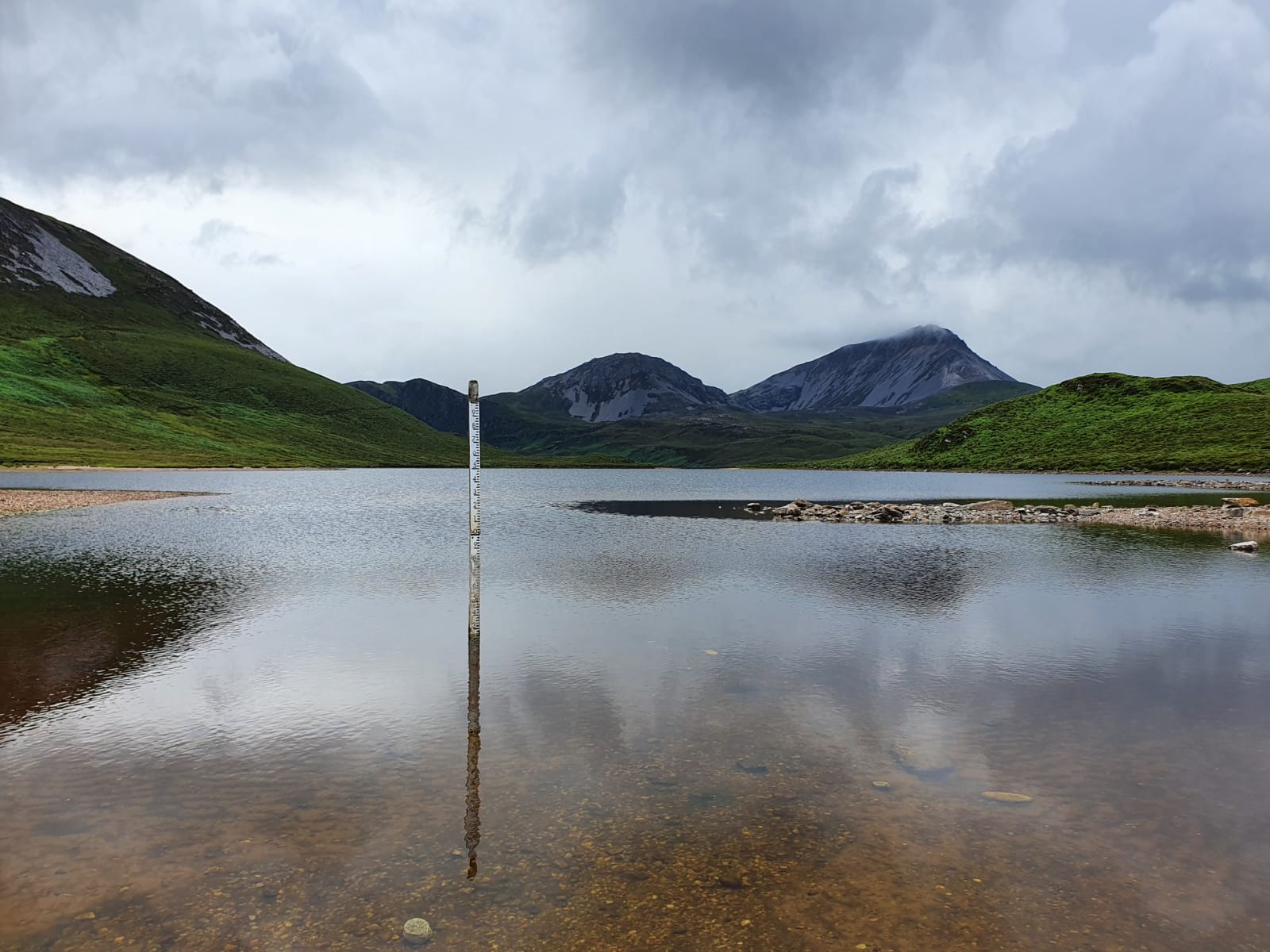 A serene landscape featuring a calm lake surrounded by green hills and mountains under a cloudy sky. In the center of the lake, there is a vertical measuring stick partially submerged in the water, indicating water levels. The reflection of the hills and sky can be seen on the surface of the lake.
