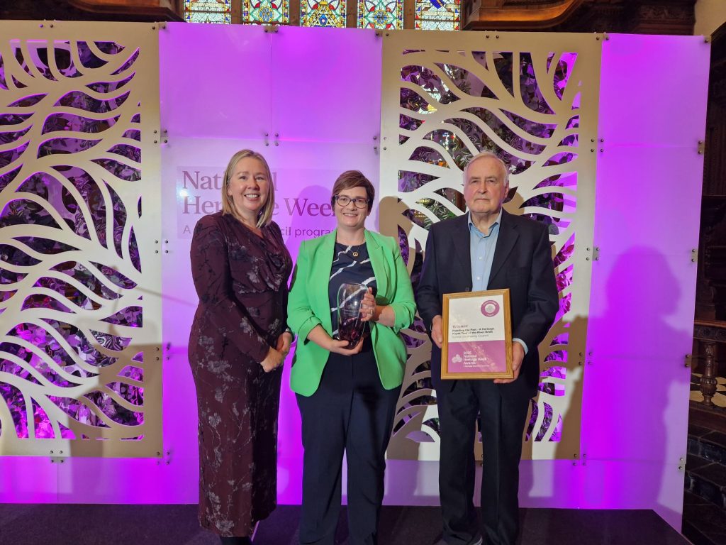 Three individuals stand on a stage during National Heritage Week. One person holds a trophy, another holds a framed certificate, with a purple-lit backdrop featuring leaf patterns behind them.
