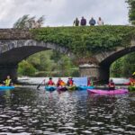 Kayakers paddle under a historic stone bridge covered in greenery, while onlookers stand above. The river is calm and surrounded by lush vegetation.