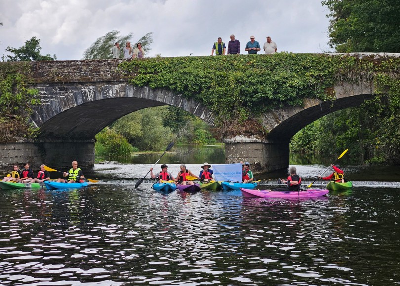 Kayakers paddle under a historic stone bridge covered in greenery, while onlookers stand above. The river is calm and surrounded by lush vegetation.