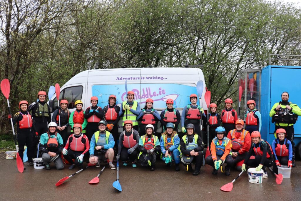 A group of people wearing helmets and life jackets holding paddles, posing in front of a van with the text “Adventure is waiting… GoPaddle.ie.” Buckets and gear are placed on the ground in front of them, with trees in the background.