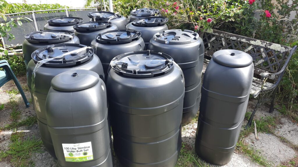 Several large black plastic water butts arranged outdoors on a paved area near a metal garden bench, with greenery and flowers in the background. A visible label on one butt reads “100 Litre Slimline Water Butt Set.