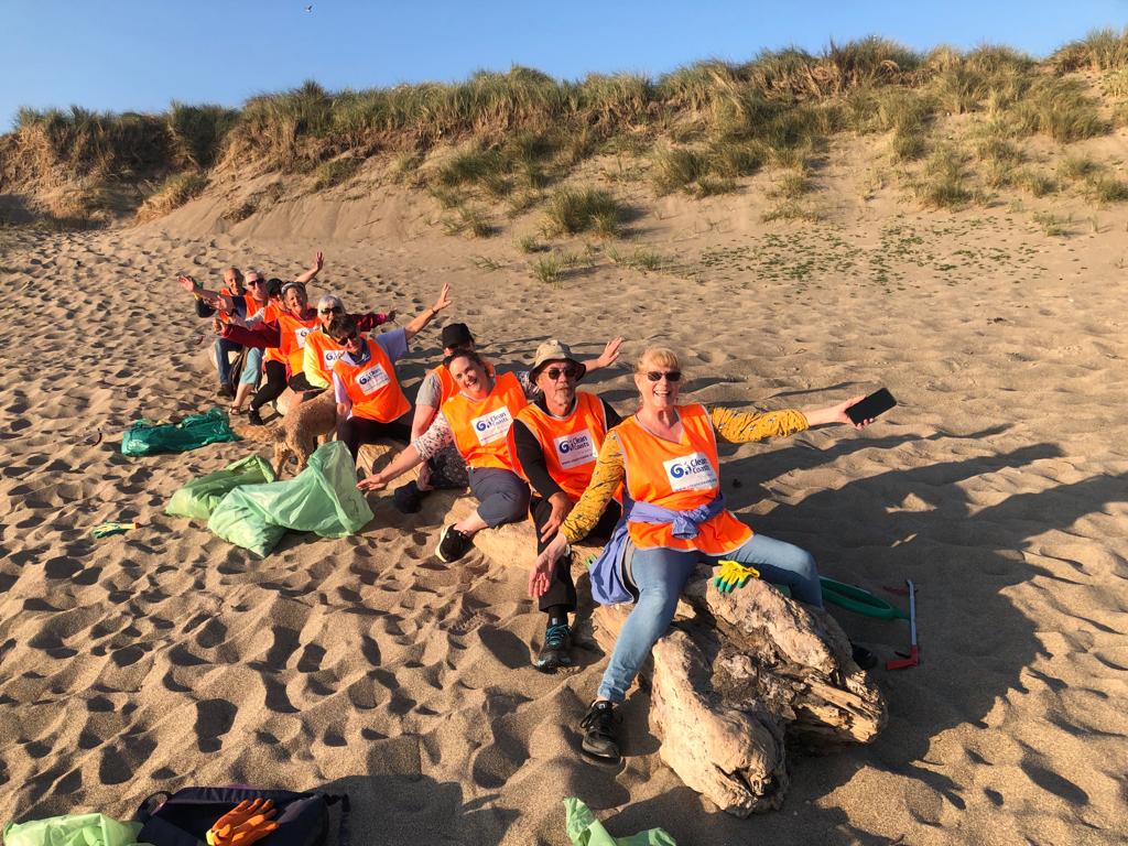 A group of people wearing bright orange vests sitting on driftwood on a sandy beach, with green litter bags and gloves nearby. Sand dunes and grass are visible in the background under clear skies.
