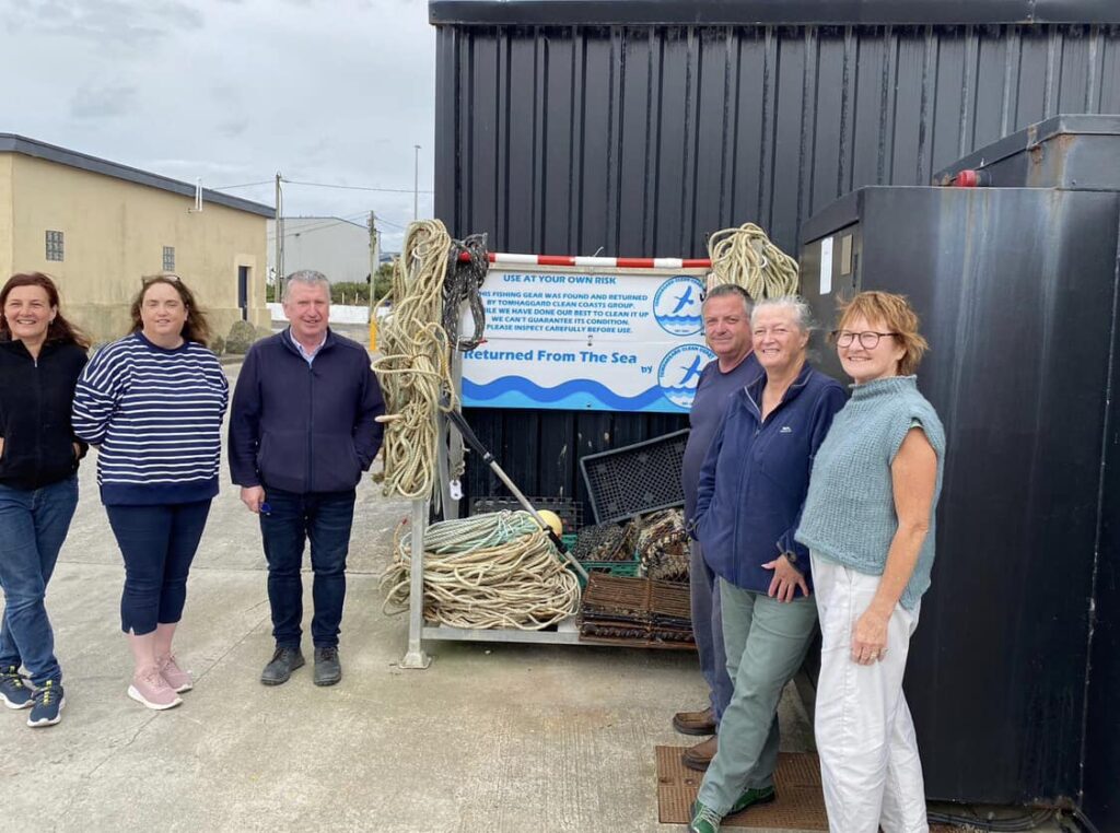 Several people standing near a black building with a sign that reads “Returned From The Sea.” Fishing ropes, nets, and equipment are stacked against the wall beneath the sign.