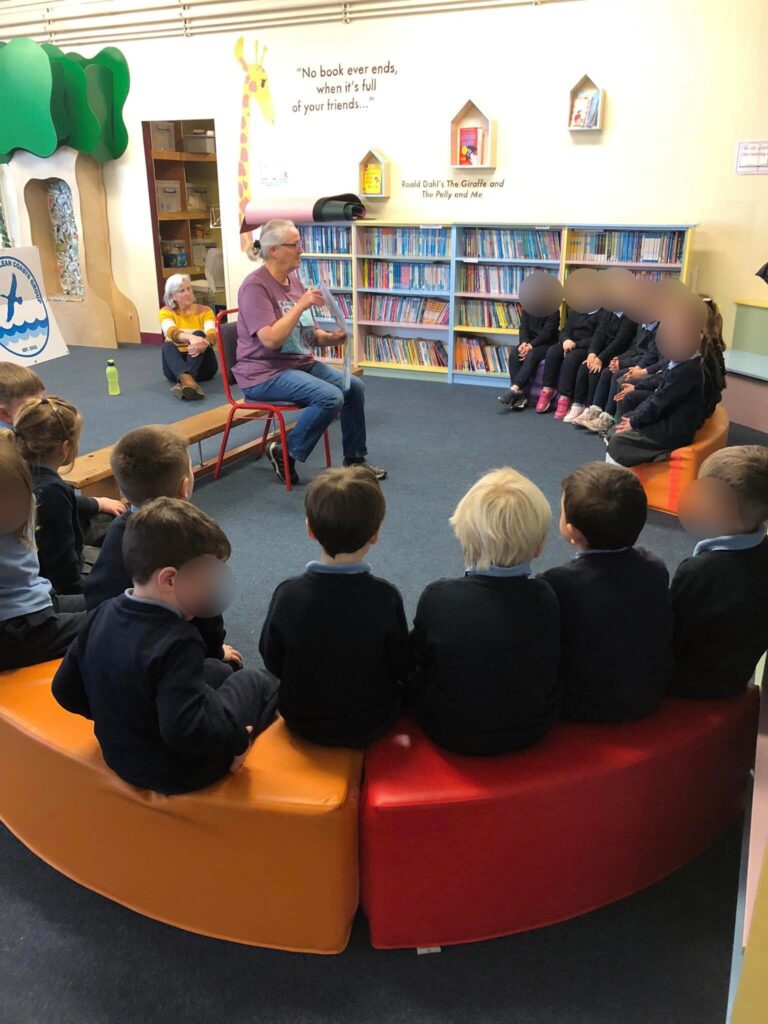 A group of young children seated on colorful curved benches in a library, listening to an adult who is sitting on a chair and speaking. Bookshelves filled with books line the wall behind, with decorative elements and quotes on the wall.