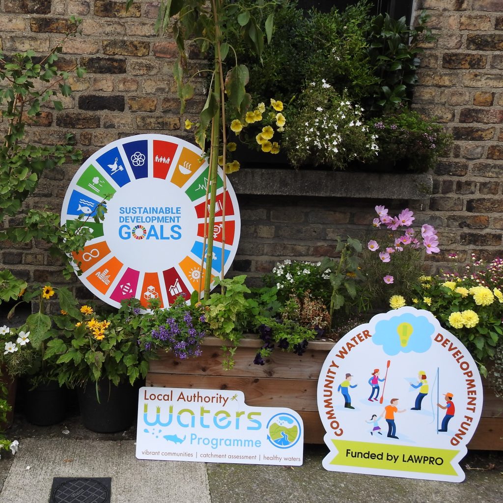 A colorful outdoor display featuring two signs among vibrant flowers and greenery against a brick wall. One sign shows the United Nations Sustainable Development Goals wheel with icons and text reading ‘Sustainable Development Goals.’ Another sign promotes the Local Authority Waters Programme with the tagline ‘vibrant communities | catchment assessment | healthy waters.’ A third sign highlights the Community Water Development Fund, funded by LAWPRO, with illustrations of people engaging in water-related activities.
