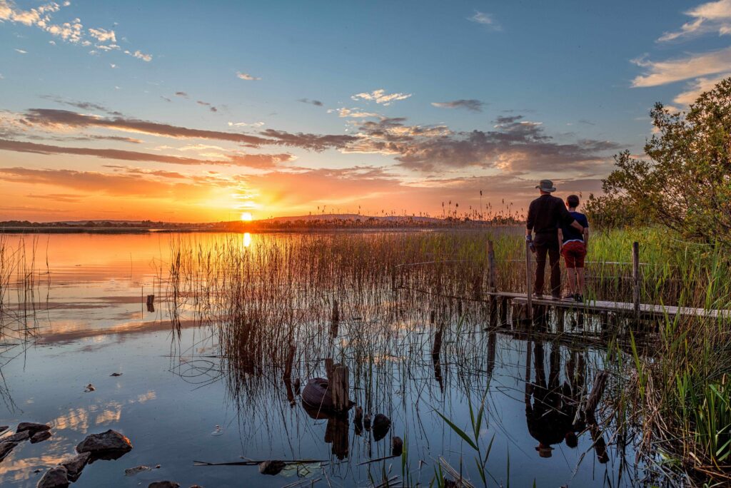 Two people standing on a wooden jetty beside a reed‑filled lakeshore at sunset. The sun is low on the horizon, casting orange and gold reflections across the calm water, with light clouds scattered in the sky.
