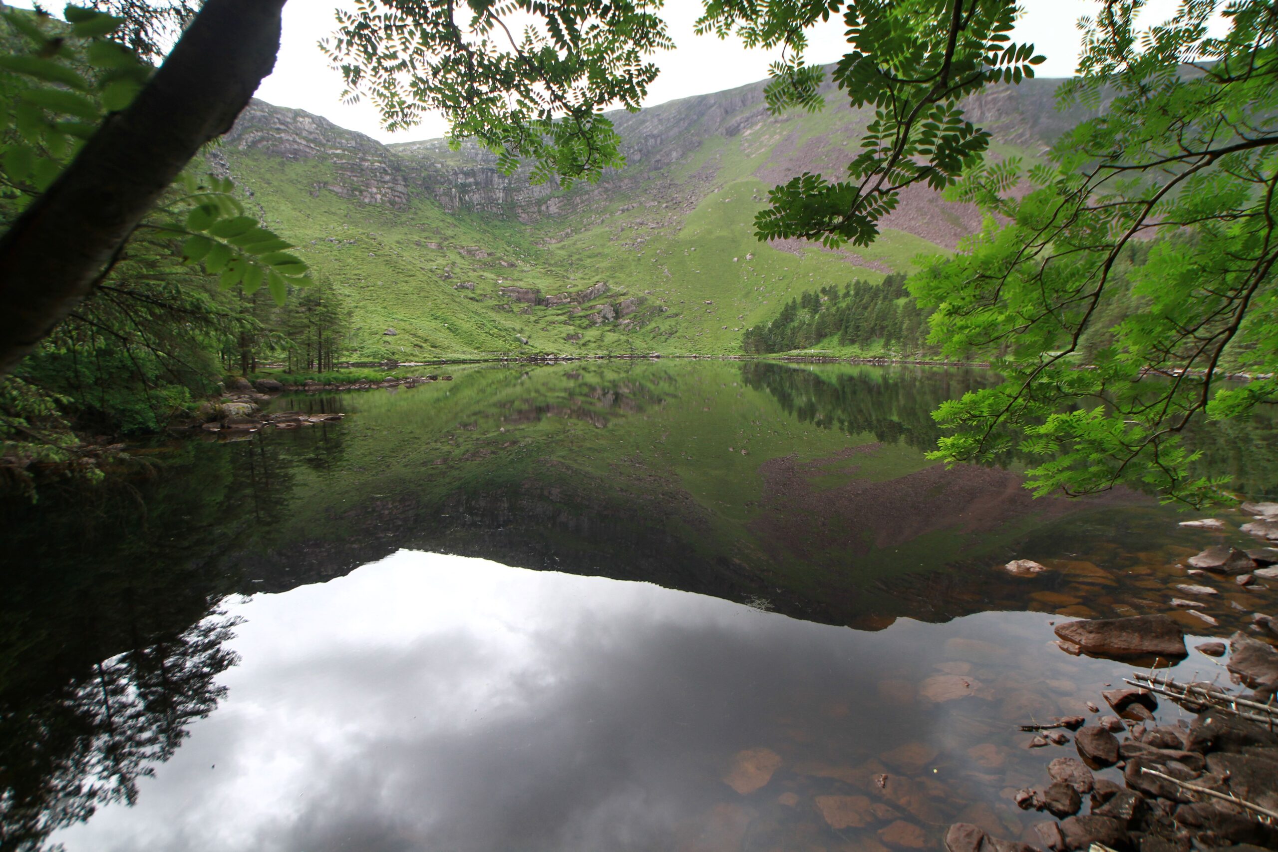 A still lake reflecting steep green mountains and a cloudy sky. Trees frame the view from both sides, and rocks are visible beneath the clear water at the lake’s edge.