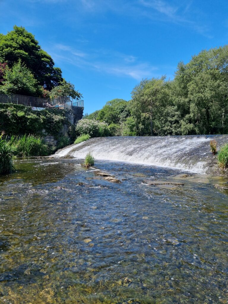 Shallow river flowing over a low stone weir, with clear water in the foreground, green trees along the banks, and a viewing platform above the left bank under a bright blue sky.