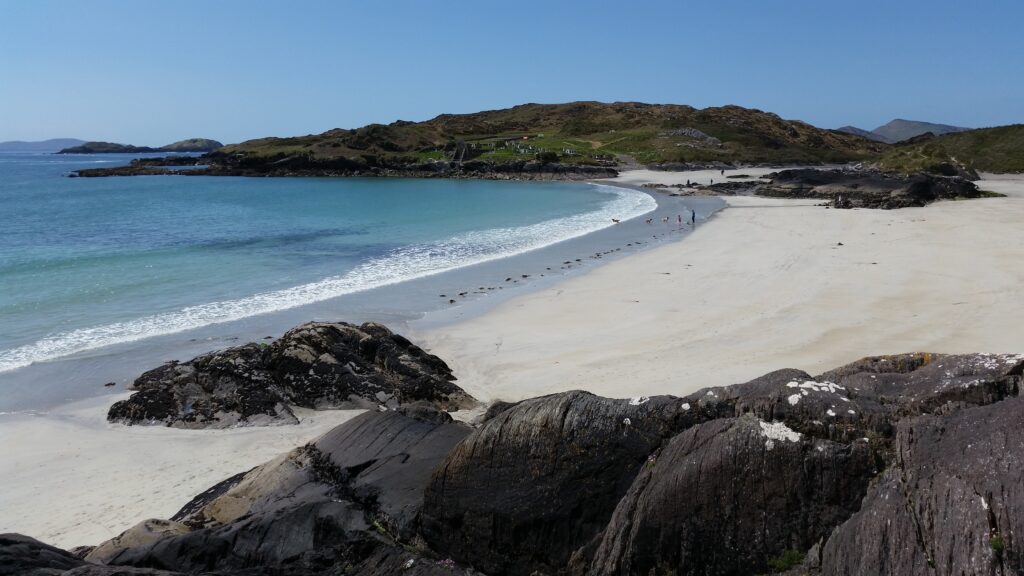 Doire Fhíonáin (Derrynane) beach in County Kerry, with pale sand curving around a sheltered bay of clear turquoise water. Rocky outcrops lie in the foreground, while grassy hills and scattered buildings rise behind the beach under a clear sky.