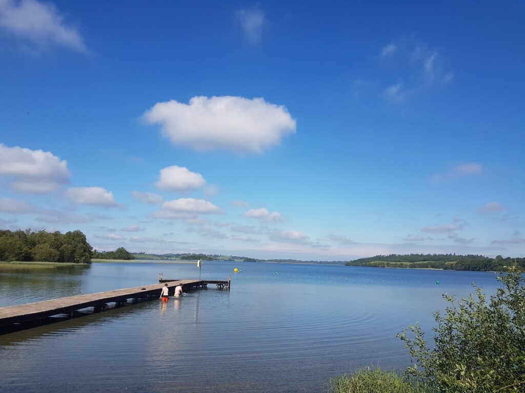 People enter the water beside a wooden swimming pier at The Cut on Lough Lene in County Westmeath. The calm lake is surrounded by low green hills and trees, with bathing marker buoys visible on the water under a bright blue sky with scattered clouds.