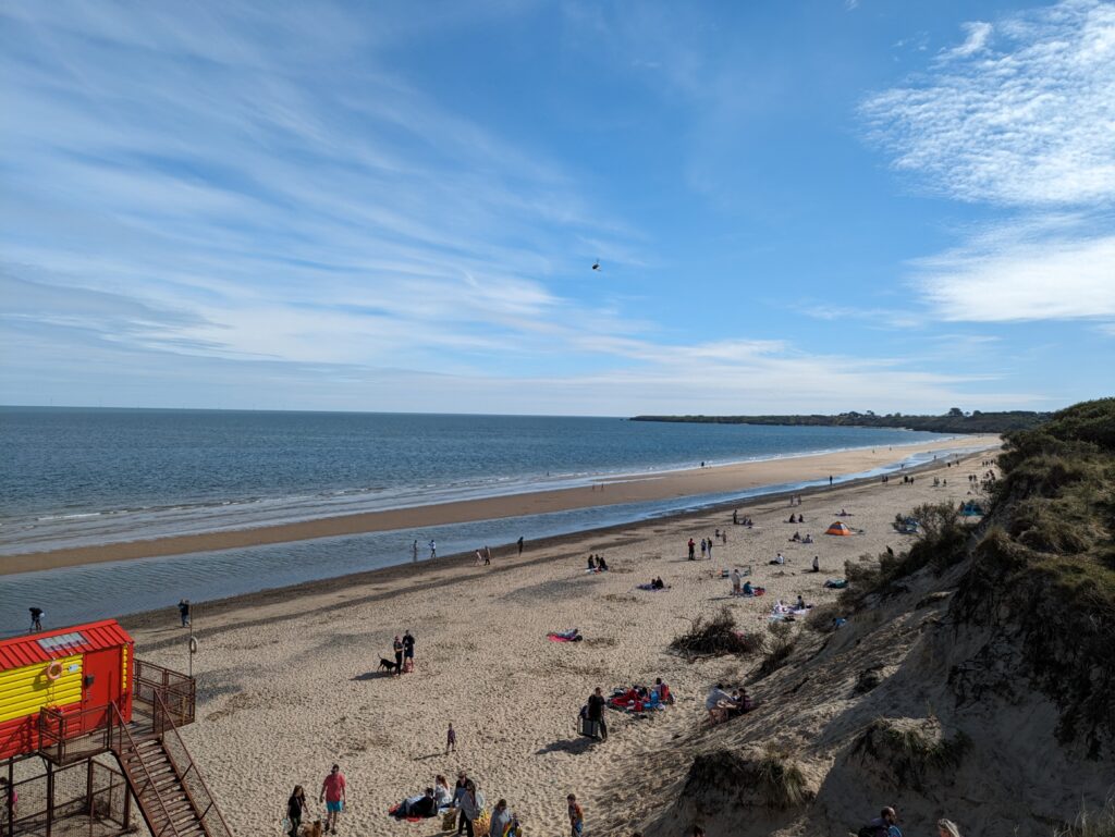 Wide view of Brittas Bay in County Wicklow, showing a long sandy beach with shallow tidal pools, the sea stretching to the horizon, and many people spaced along the shore. Sand dunes rise on the right, and a lifeguard structure appears in the foreground beneath a clear blue sky.